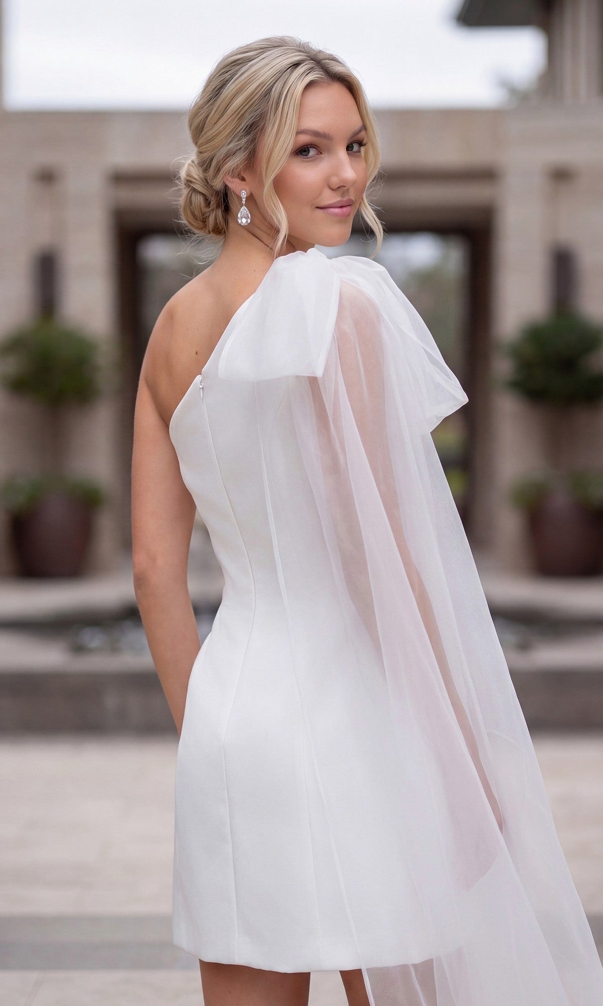 A woman wearing the Nox Anabel Y7003W White Mini Hoco Dress, featuring a one-shoulder design and sheer flowing overlay, stands outdoors and looks back over her shoulder.